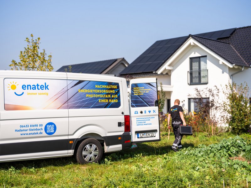 Ein Transporter mit enatek-Branding steht mit geöffneten Hecktüren vor einem Einfamilienhaus. Ein Mitarbeiter räumt Werkzeuge aus dem Transporter und bringt sie zum Haus.