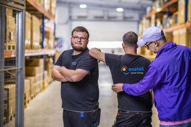 Das Foto zeigt zwei Mitarbeitende von enatek, einer schaut in die Kamera, der andere wendet der Kamera den Rücken zu). Eine weitere Person glättet die Falten der Arbeitskleidung.