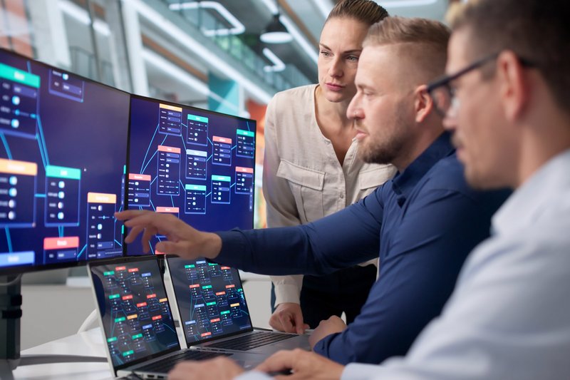 Drei Menschen arbeiten an mehreren Monitoren und sehen auf ein Prozessschaubild.