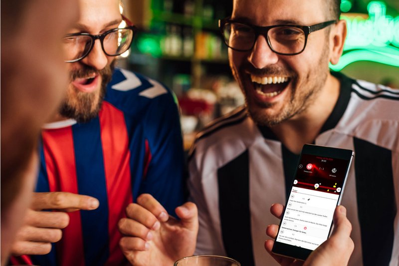 Drei Fußballfreunde feiern die Spielentwicklung der Fortuna Köln durch Verfolgung des Liveticker auf dem Smartphone