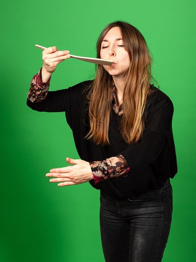 Kathrin Scholz umfasst mit dem linken Arm einen imaginären Topf und kostet an einem Löffel in der rechten Hand